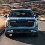 Front grille view of a blue 2027 Kia Carnival driving a winding road.