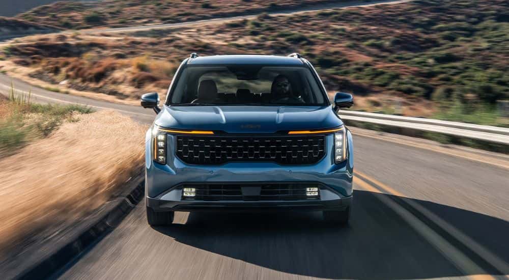 Front grille view of a blue 2027 Kia Carnival driving a winding road.