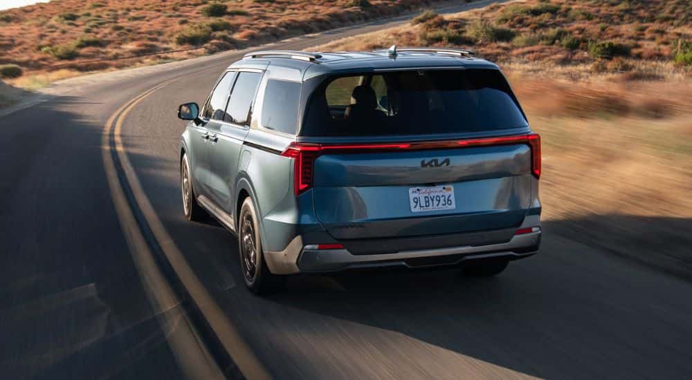 Rear view of a blue 2027 Kia Carnival SX Prestige driving a winding road.