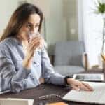 Young business woman working at home and drink glass of water. She drinking water while working at the laptop computer. Brown hair busy freelancer woman working at computer and connecting to the internet