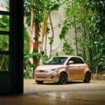 A 2024 Fiat 500e is parked in front of a building, showcasing its vibrant color against the urban backdrop.