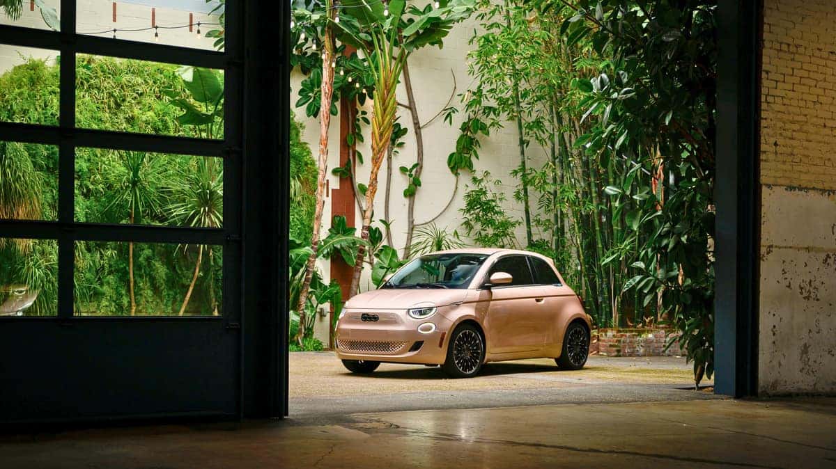A 2024 Fiat 500e is parked in front of a building, showcasing its vibrant color against the urban backdrop.